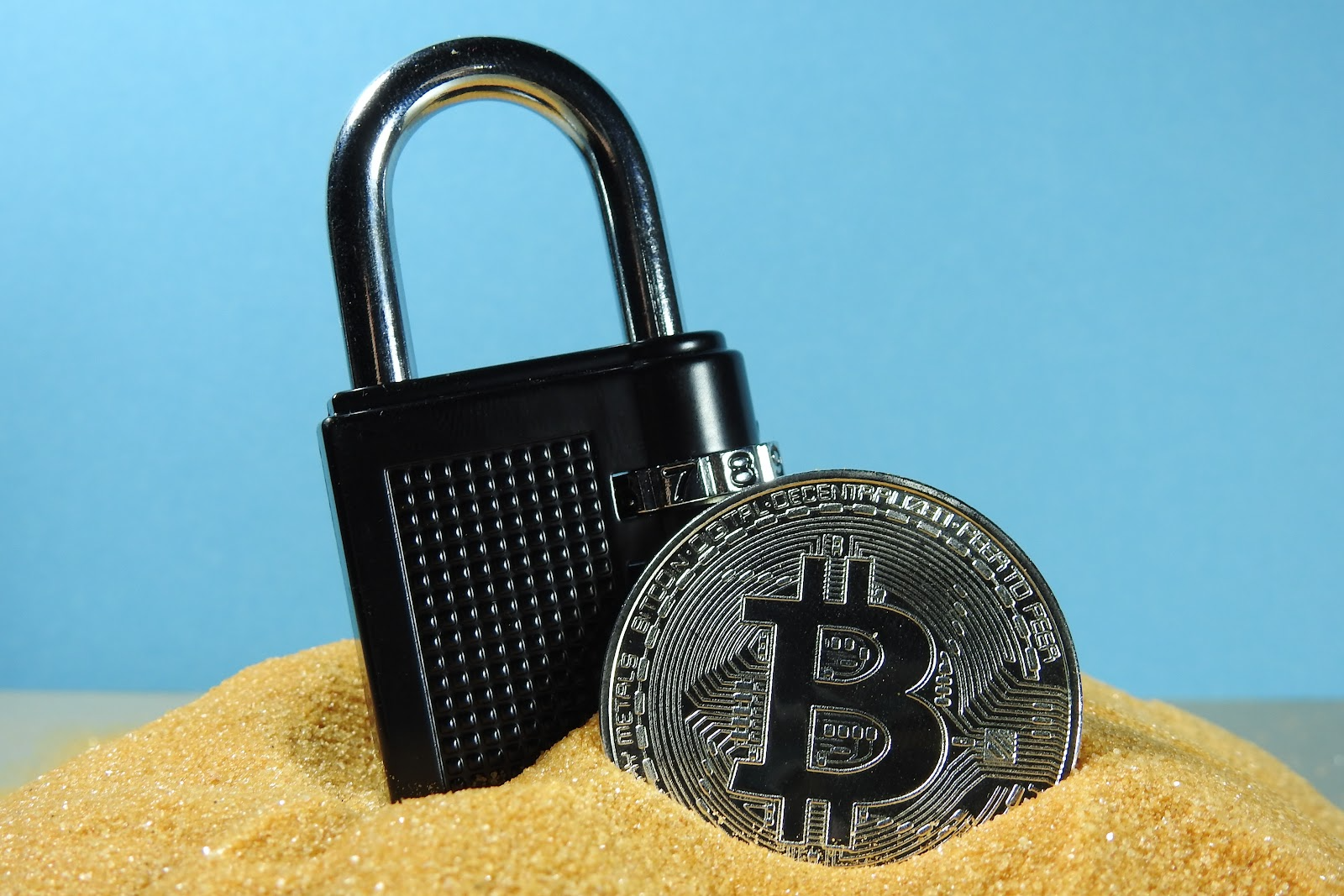 a padlock and a bitcoin on sand in front of a blue background